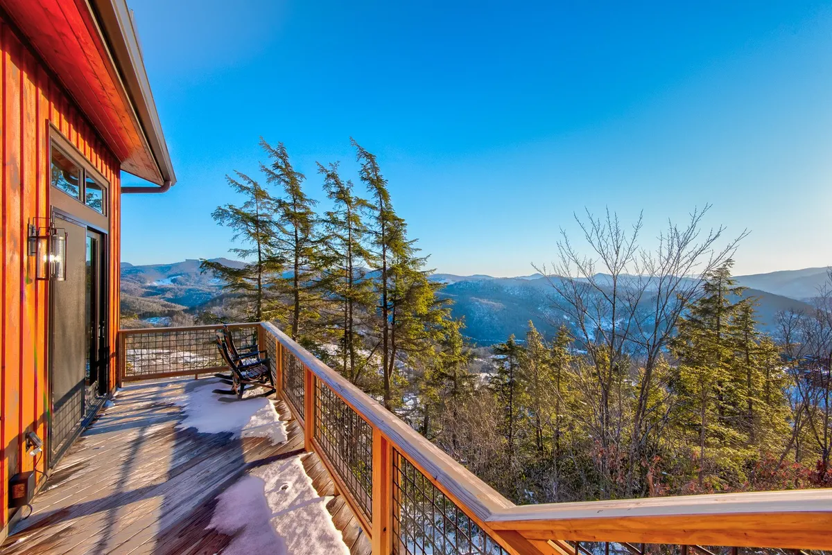 This image features a peaceful outdoor deck with a wooden railing and a rocking chair, overlooking expansive mountain views and surrounded by trees. The scene invites visitors to relax and soak in the natural beauty just outside the home.