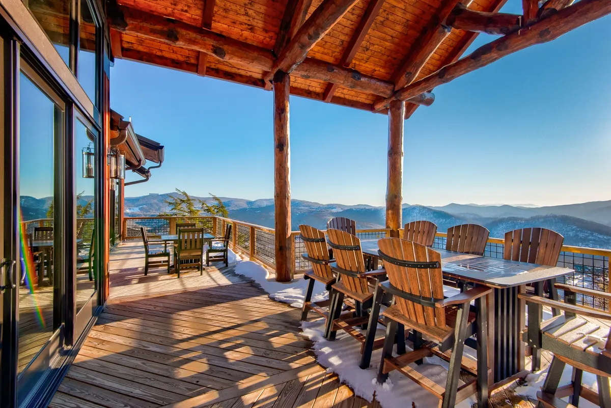 A tranquil covered deck is displayed here, with comfortable outdoor seating, a wooden dining table, and a glass railing framing the sweeping long-range mountain views. The space is designed for al fresco dining and relaxation, connecting the interiors to breathtaking outdoor scenery.