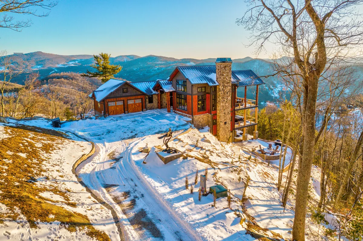 The modern house stands amidst a snow-covered landscape, its stone chimney and wooden sculptures adding rustic charm. Soft evening light hints at cozy nights inside, while the deck and surrounding snow conjure images of winter activities and gatherings.
