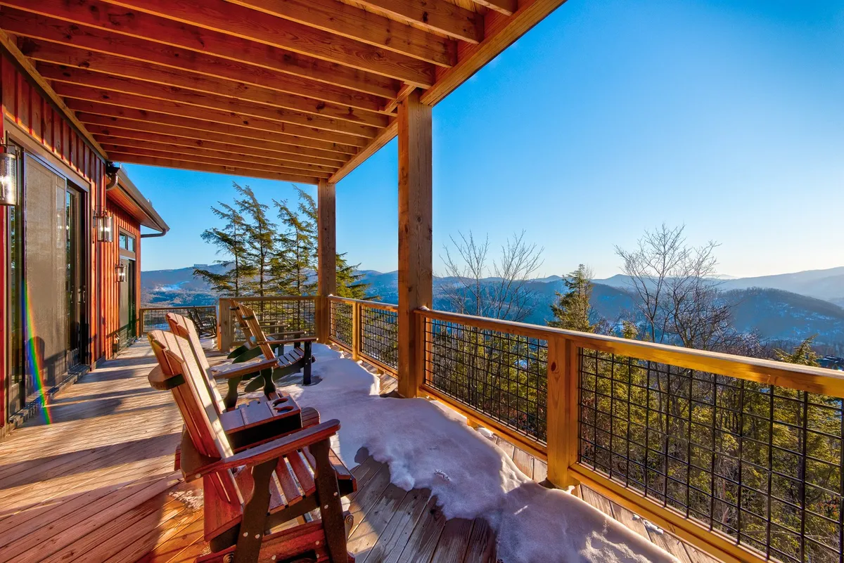 A wooden deck furnished with classic rocking chairs overlooks sweeping mountain vistas. Patches of snow highlight seasonal transitions, while the scene is framed by natural wood and open sky, inviting visitors to experience the outdoors year-round.
