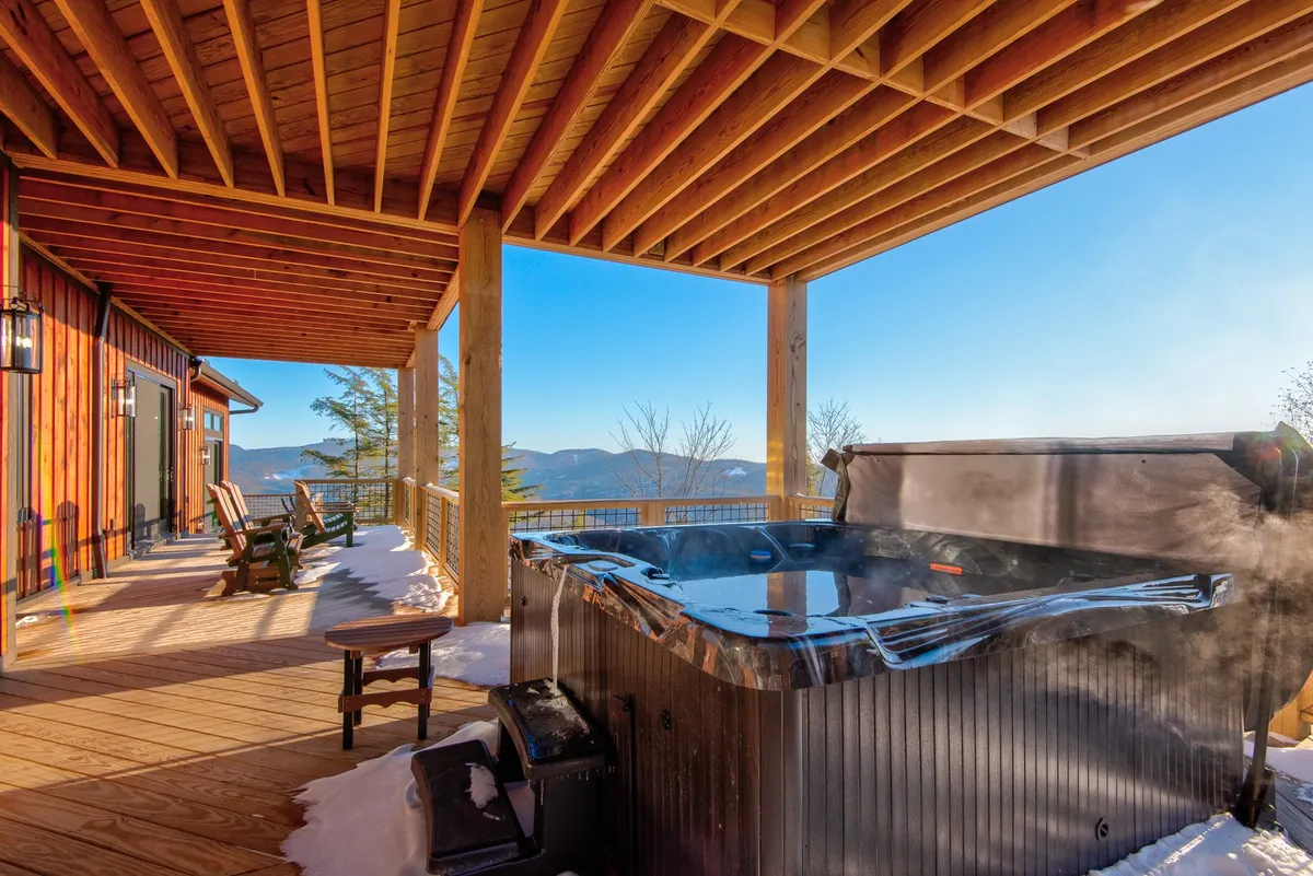 An exterior shot of the lower deck, highlighting a bubbling hot tub with scenic mountain views. Wooden rocking chairs flank the hot tub, creating a perfect setting for relaxation and conversation, all framed by the natural beauty of the outdoors.