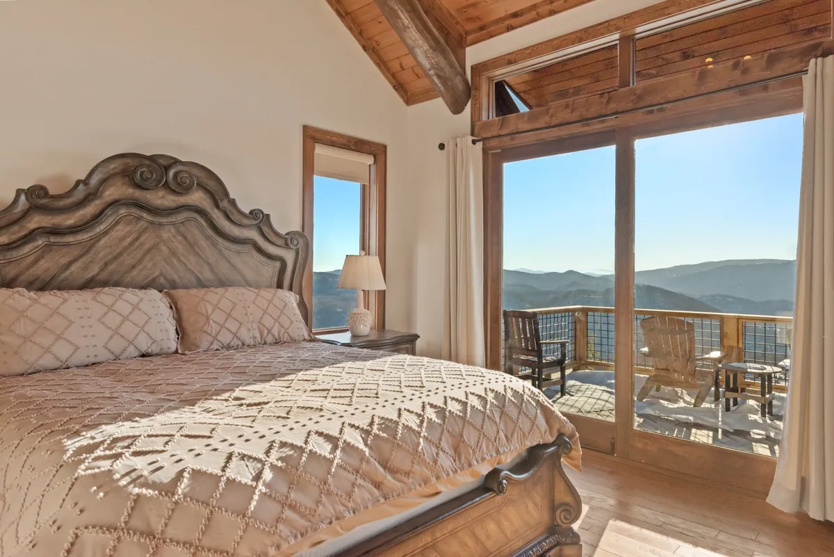 An elegant bedroom with an ornate king bed, large windows framing panoramic mountain views, and access to a wooden balcony. The space is serene, welcoming, and exemplifies a private retreat.