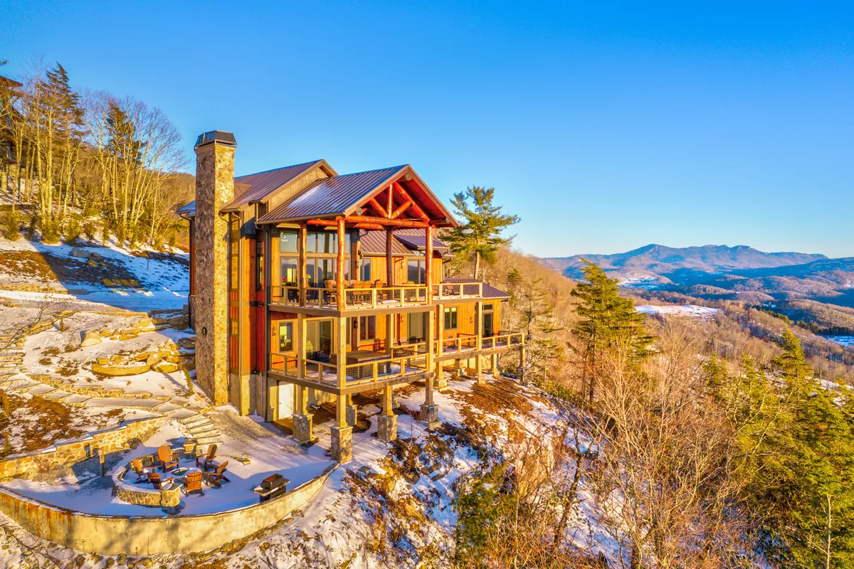 The image features a grand wooden mountain home with a striking stone chimney, surrounded by a spacious outdoor seating area. The home is perched to capture stunning panoramic mountain views, perfect for savoring sunsets, relaxing by the fire pit, or sharing unforgettable moments with loved ones. The atmosphere is tranquil, inviting guests to unwind and reconnect in nature's embrace.