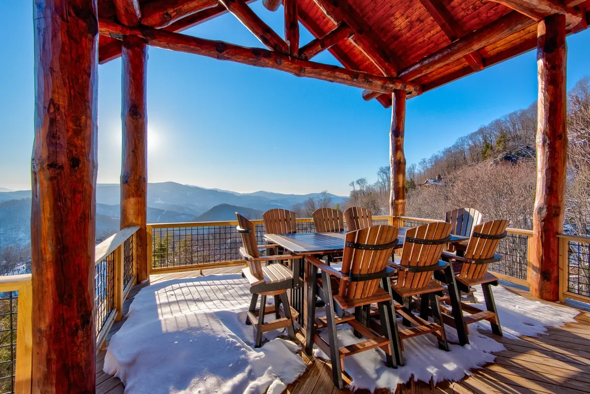 An inviting exterior scene shows a large wooden dining table and surrounding chairs positioned on a deck, all set against a backdrop of sunlit mountain scenery. The area is ideal for group gatherings and outdoor meals, with ample space for camaraderie and enjoyment of the natural setting.