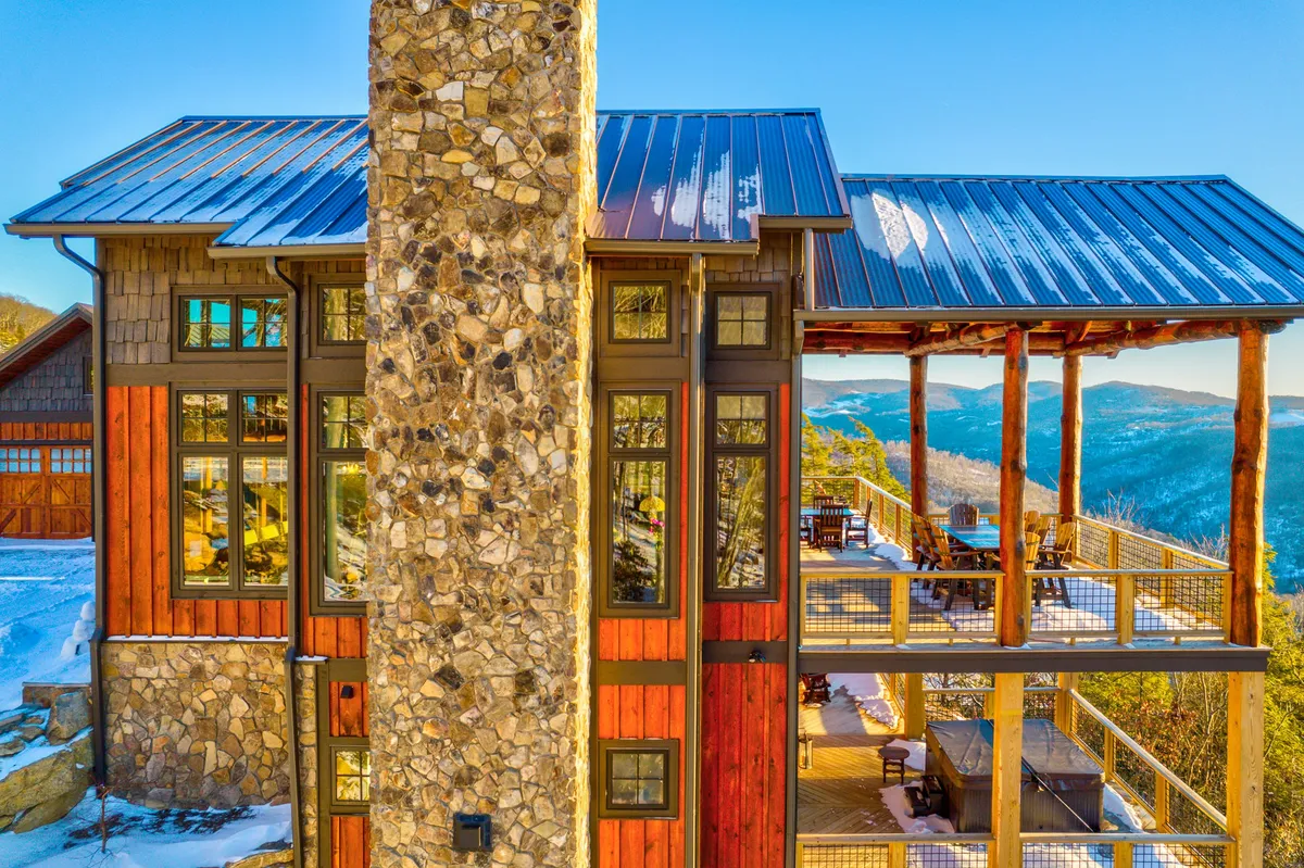 An exterior view of the cabin showing its stone chimney, expansive windows, and a large wooden deck. The deck appears ready for relaxation and entertaining, with panoramic mountain vistas and the welcoming ambiance of mountain living.