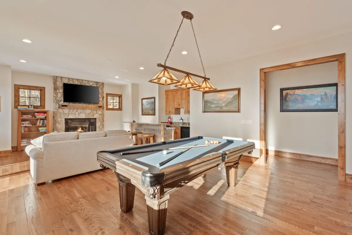 This image captures a lively entertainment den featuring a pool table as the central focus, with a cozy seating area by a fireplace and a kitchen area in the background. The room is designed for both relaxation and recreation, evident from the comfortable sofa and the inviting fireplace, while the pool table sets the stage for friendly competition.