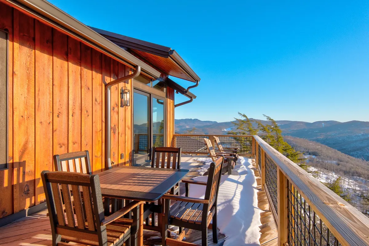 A spacious wooden deck set against a backdrop of snow-covered mountains, featuring an outdoor dining table and comfortable seating. The deck's railing frames the sweeping mountain views, inviting guests to relax and appreciate the natural beauty in any season.