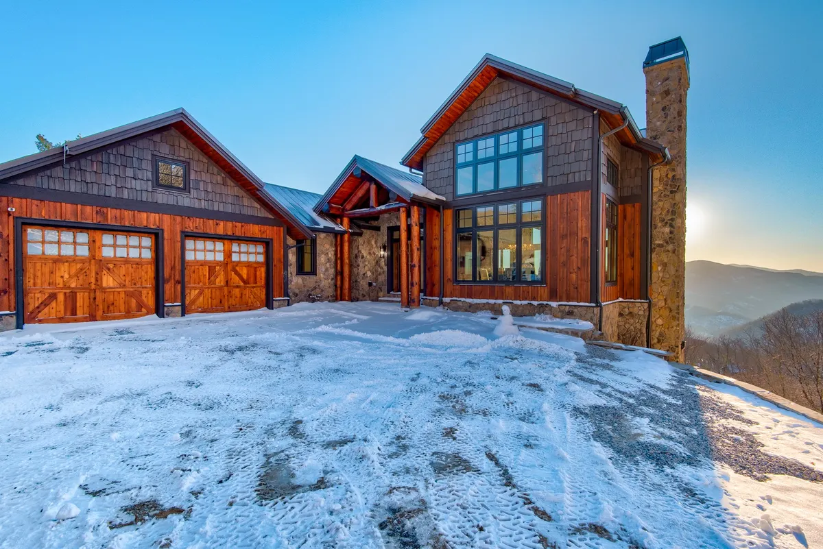 A view of the home's wooden garage doors, stone chimney, and large glass windows. The snow-covered driveway hints at easy winter access and practical amenities for storing gear, while still showcasing the home’s inviting, mountain aesthetic.
