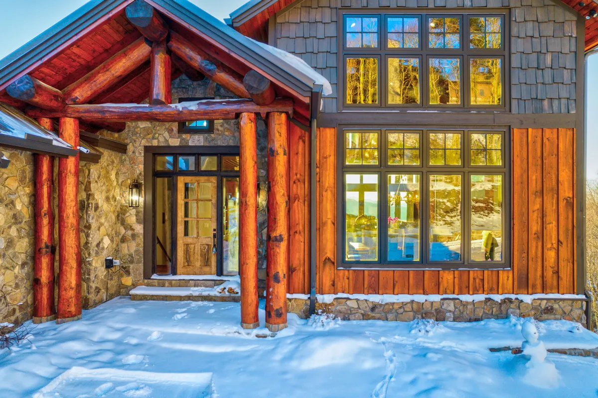 The front exterior of a welcoming home, featuring large windows, wooden pillars, and stone walls. Snow dusts the ground, highlighting the home’s warmth and inviting nature. The scene suggests both year-round appeal and a gateway to the community’s amenities.