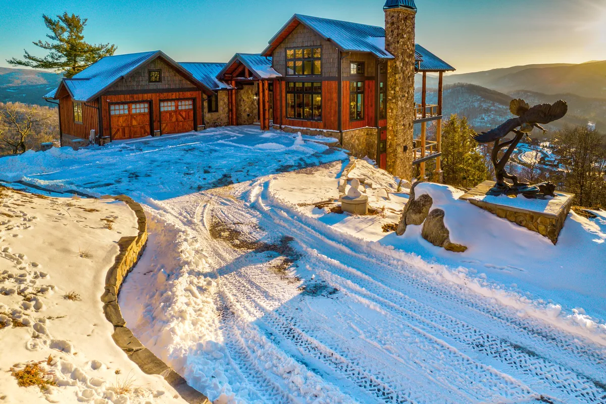 This exterior shot captures the house with a snow-covered driveway and a distinctive sculpture in the foreground, surrounded by a tranquil winter landscape. The scene highlights the home's entry point and its placement within the community environment.